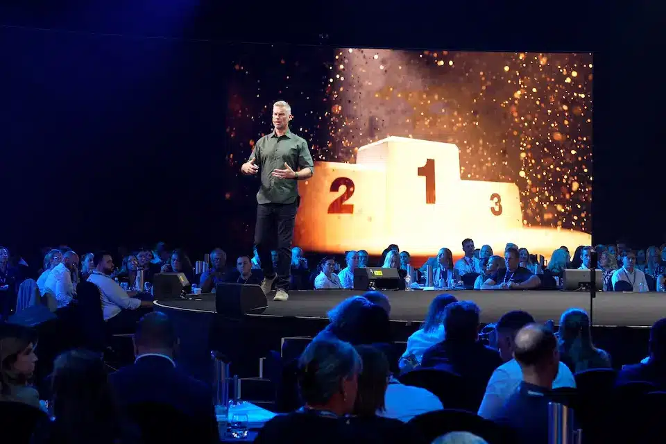 Mark Dobson presenting to packed audience in dark room with illuminated podium image on big screen behind him